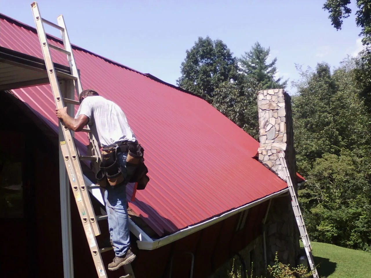 Roofer on a ladder beside a red metal roof and stone chimney — Soffit & Fascia Repair by Roofing Brothers in Downey